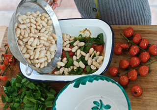 Pouring beans into a container of Bean Salad, tomatoes and herbs on the table beside it