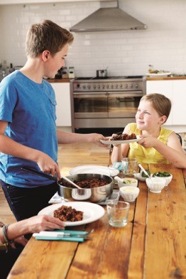 Children sitting around a table, one child spooning out a meal onto a dish and handing it to a younger child