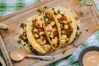 5 healthy lentil tacos fanned out on a wood board, with a spoon of sauce placed beside them