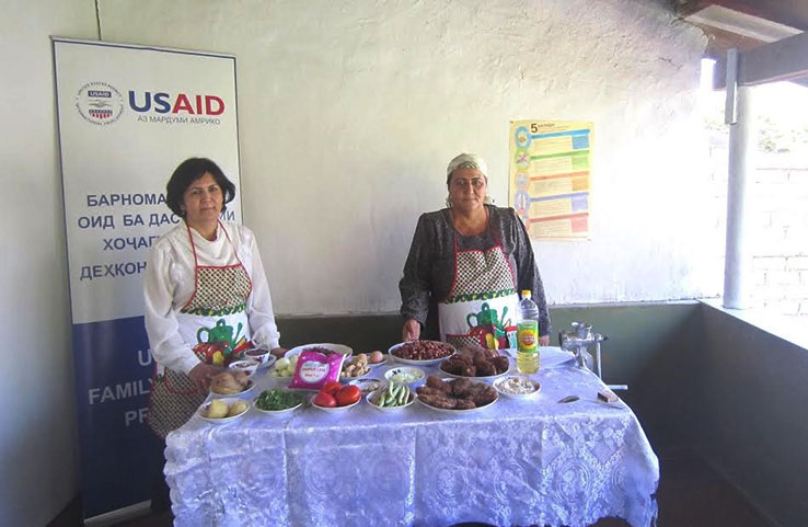 Two ladies standing behind a table of food and a US AID poster for a recipe book they helped develop